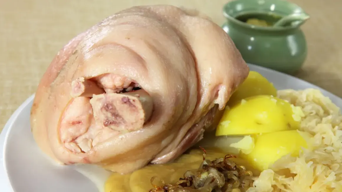 Ein deftiges, gekochtes Eisbein mit Salzkartoffeln, Sauerkraut und einer kleinen Schale mit Soße.