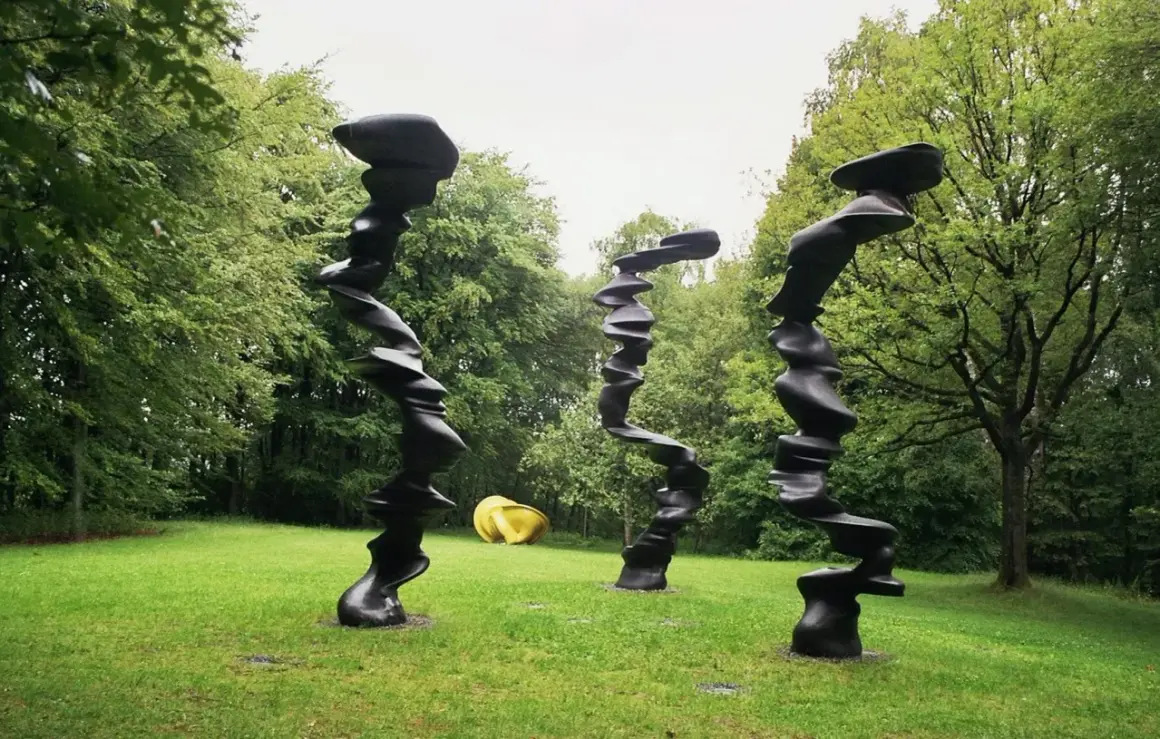 Drei schwarze, verdrehte Skulpturen stehen im **Skulpturenpark Wuppertal** auf einer grünen Wiese vor Bäumen. Eine gelbe Skulptur liegt im Hintergrund.