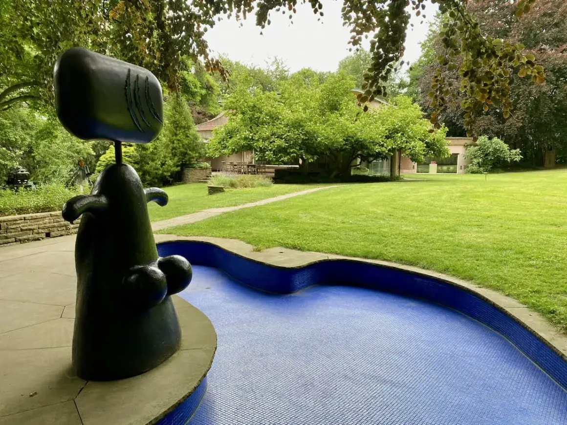 Eine abstrakte schwarze Skulptur steht neben einem blauen Mosaikbecken in einem **Skulpturenpark**. Im Hintergrund ein gepflegter Rasen und Bäume.