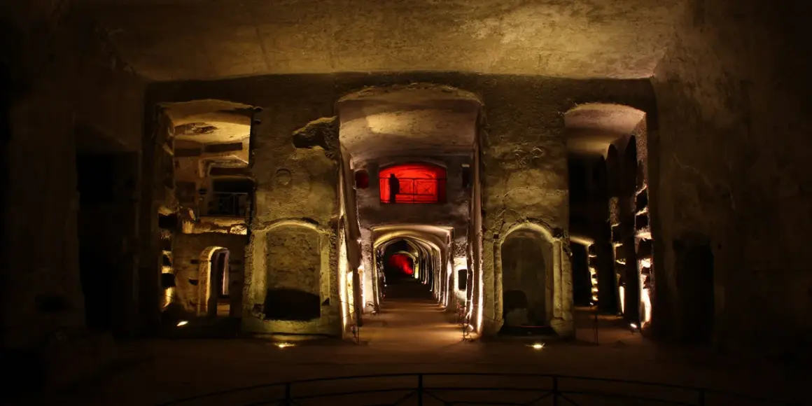 Corridoio sotterraneo illuminato, con nicchie scavate nella roccia e un'apertura rossa in lontananza, evocando l'atmosfera della tomba di San Gennaro.
