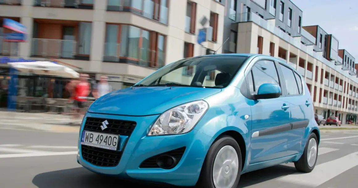 Błękitny Suzuki Splash jedzie ulicą miasta. W tle nowoczesne budynki i ludzie.