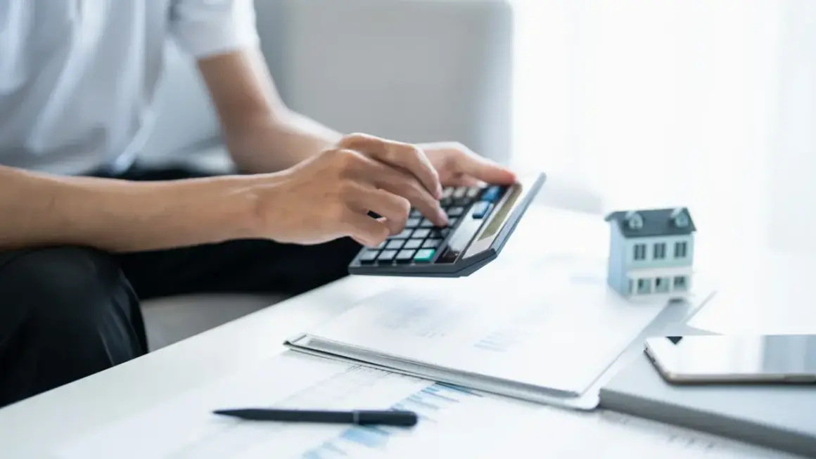 Man calculando la viabilidad hipoteca con calculadora, papeles y maqueta de casa.