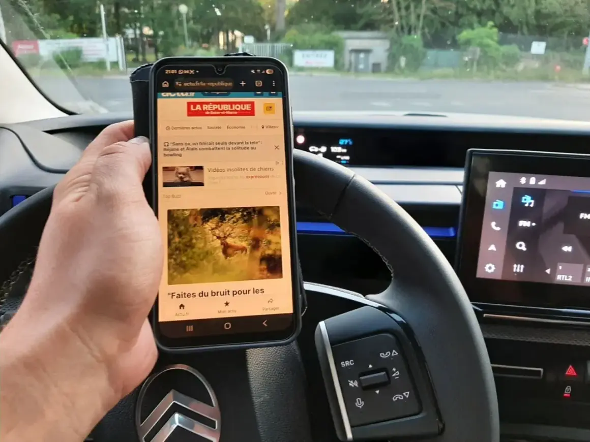 Main tenant un téléphone au volant, risque d'amende. L'écran affiche des articles d'actualité, un danger pour la conduite.
