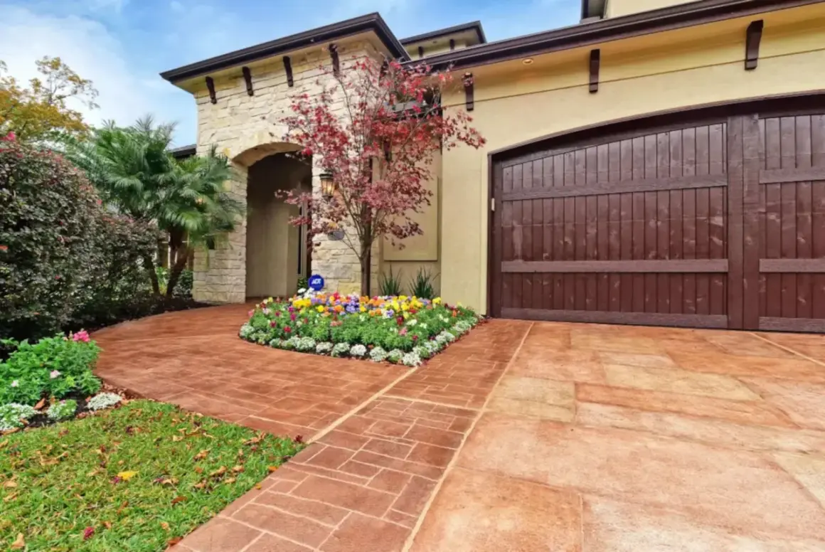 Stunning paver walkway ideas featuring a stamped concrete path leading to a home with a stone facade and a rich wooden garage door.