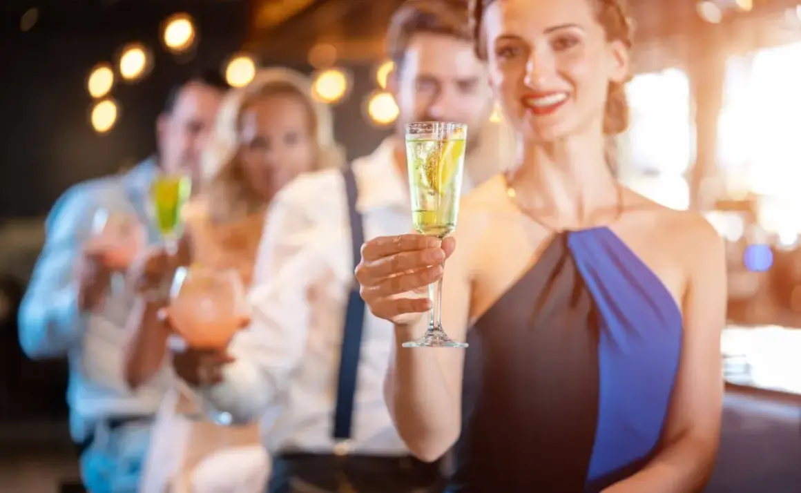 Uśmiechnięta kobieta trzyma kieliszek z drinkiem z prosecco i wódką, w tle bawią się inni goście.