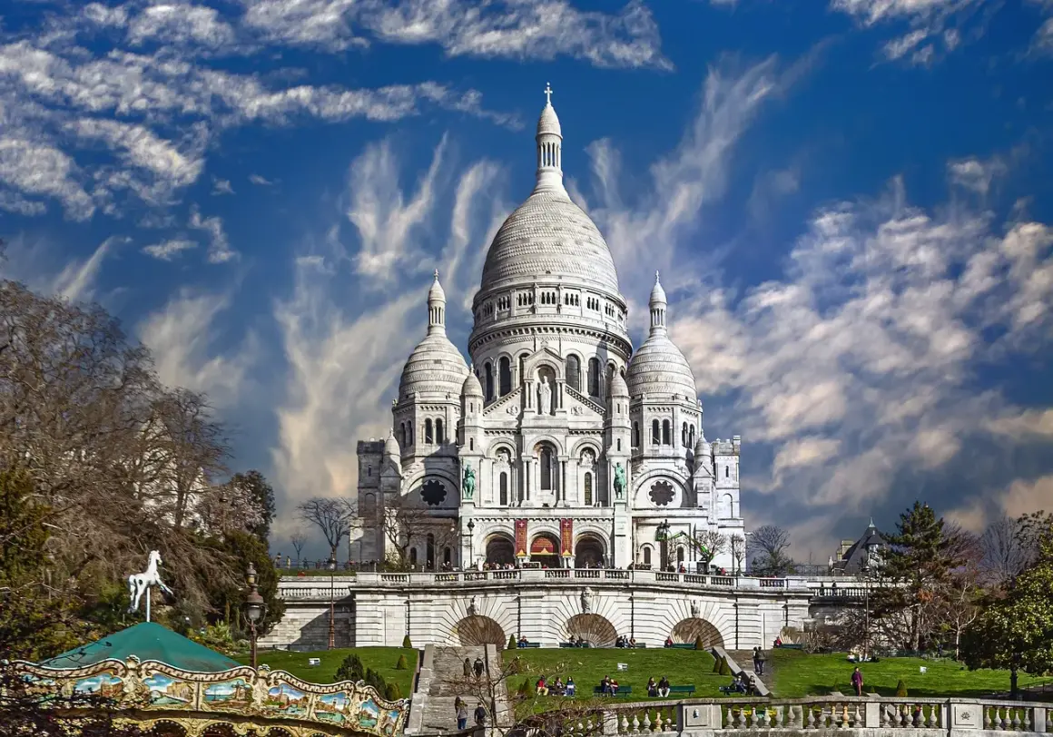 Bazylika Sacré-Cœur na wzgórzu Montmartre w Paryżu, z karuzelą w tle i dramatycznym niebem.