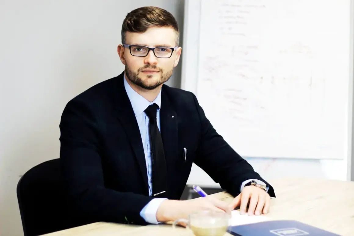 Mężczyzna w garniturze i okularach siedzi przy biurku, pisząc coś na kartce. Obok stoi filiżanka kawy.
