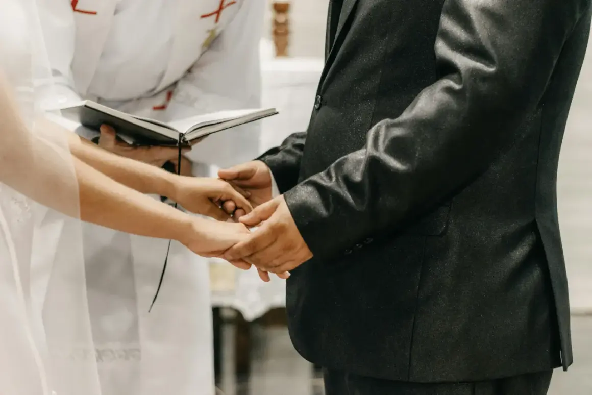 Mani che si stringono durante la dichiarazione matrimonio, con un libro aperto e un sacerdote sullo sfondo.