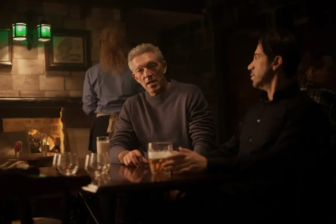Besetzung von Damaged: Vincent Cassel und Reda Kateb sitzen in einer dunklen Bar bei Bier.