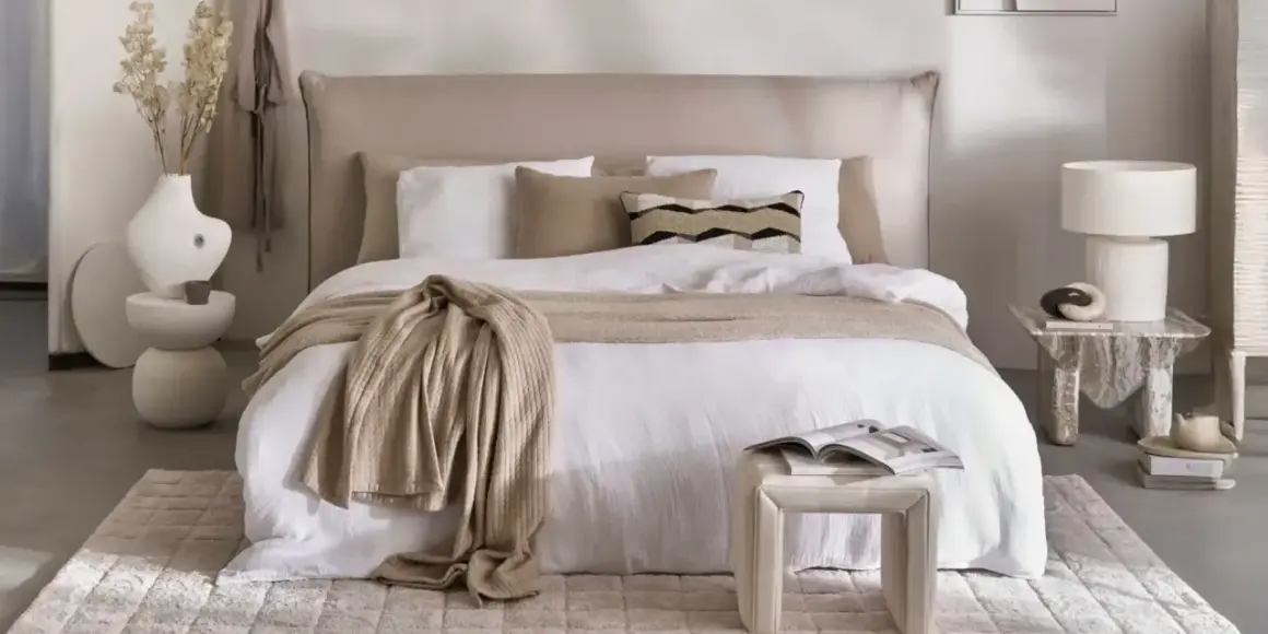 Ein gemütliches Schlafzimmer im shabby stil mit einem weißen Bett, beige Decke und Kissen. Ein Beistelltisch mit Lampe und Deko.