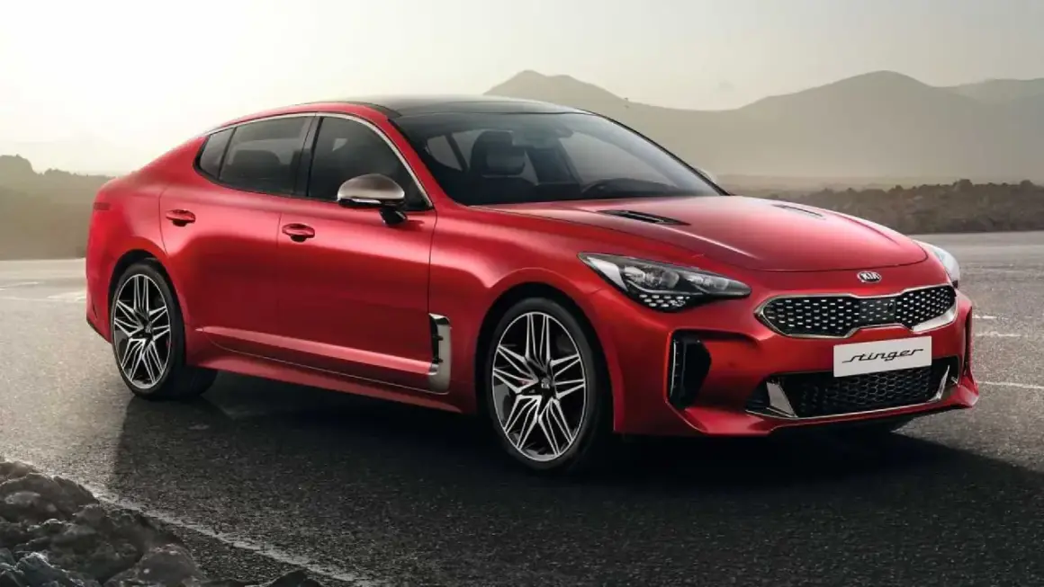 Kia Stinger CV rojo brillante, con llantas deportivas, aparcado en una carretera mojada con montañas al fondo.