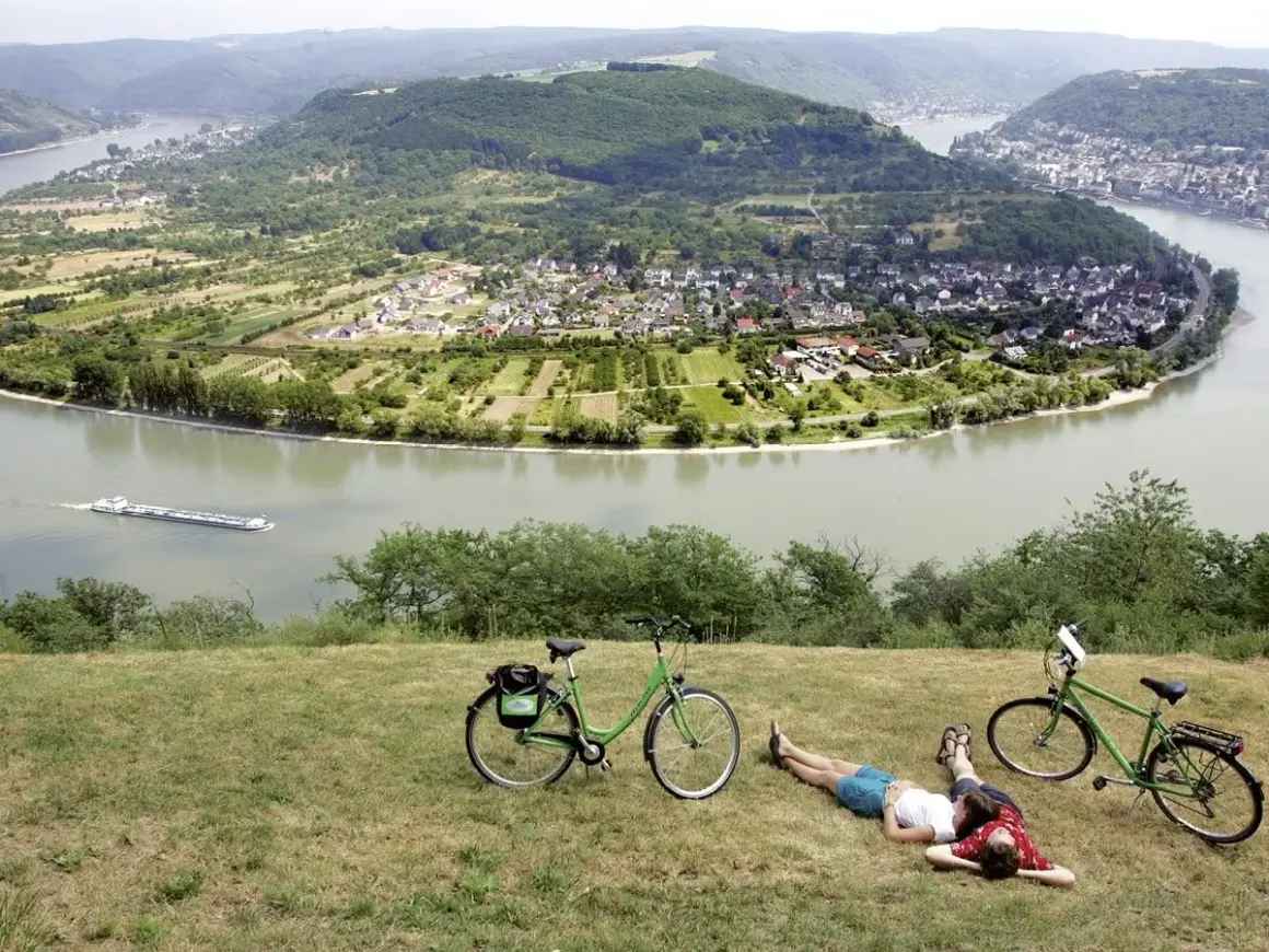 Pareja descansa junto a sus bicicletas en la ruta EuroVelo 15, con un río y un pueblo en el valle.