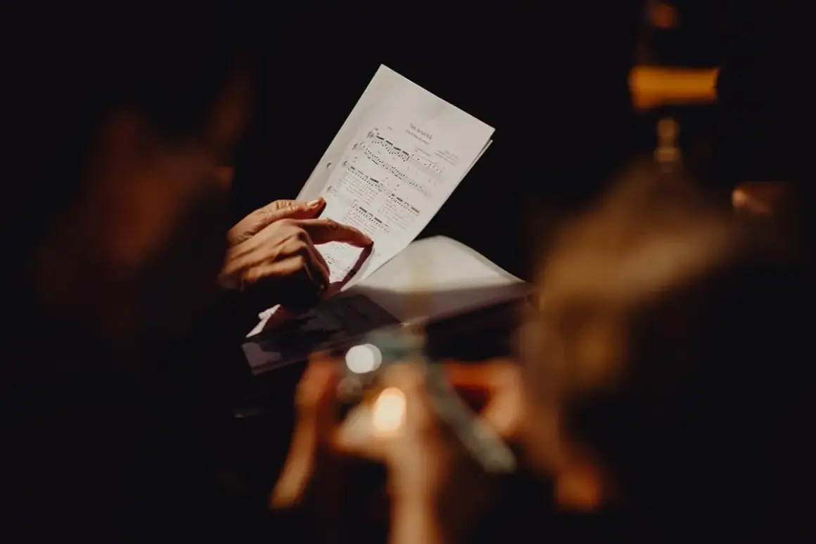 Mani che seguono le note di una canzone funerale su uno spartito, in un'atmosfera raccolta e commossa.