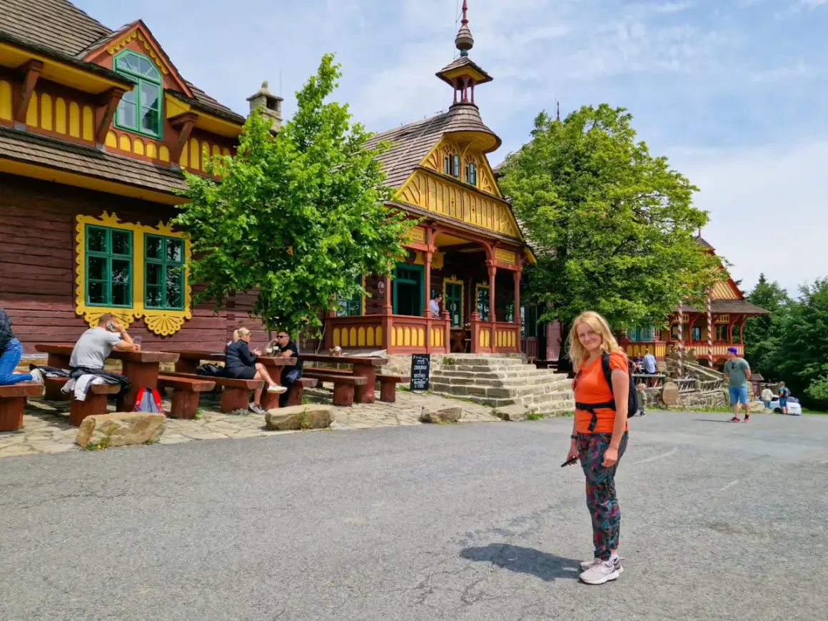 Kolorowe, drewniane budynki w Czechach, pełne ciekawostek dla dzieci. Kobieta w pomarańczowej bluzce pozuje na tle malowniczej architektury.