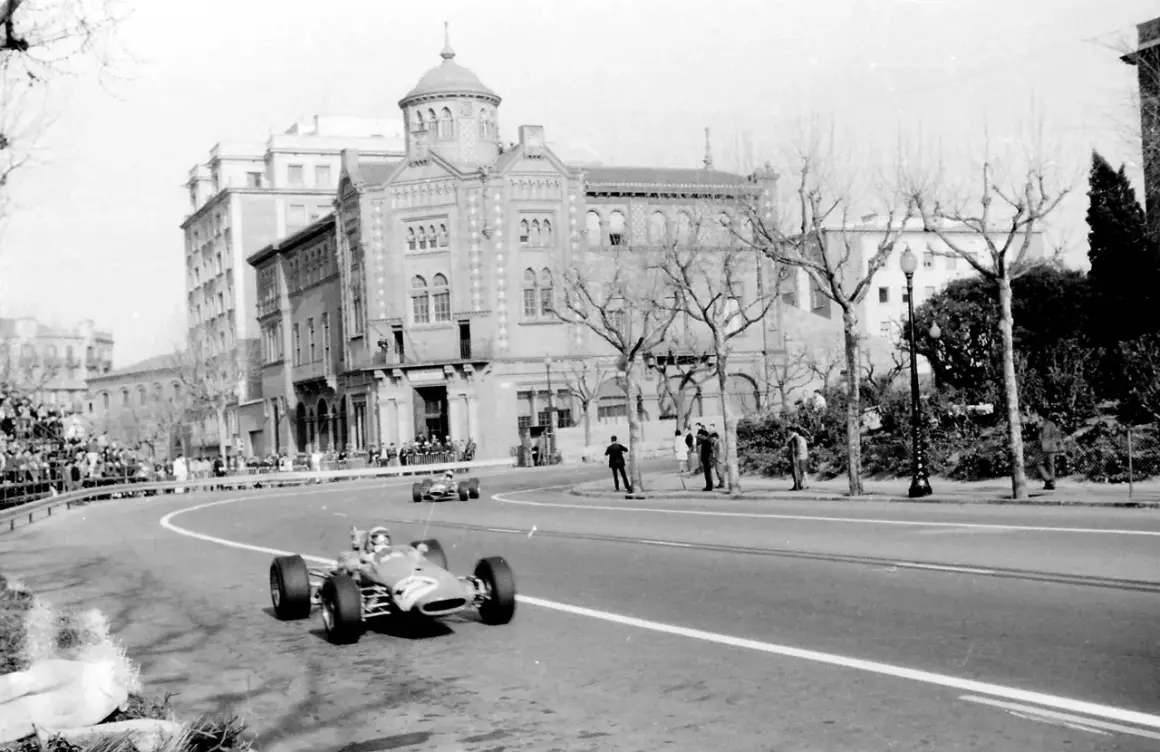 Coches de carreras compiten en el circuito de Montjuïc, con espectadores en las gradas y edificios históricos al fondo.