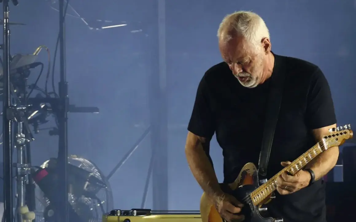 David Gilmour gra na gitarze elektrycznej na tle sceny z dymem i sprzętem muzycznym.