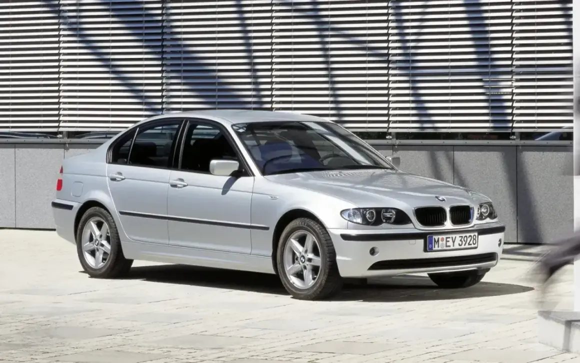 Srebrne BMW E46 sedan z charakterystycznymi podwójnymi reflektorami i grillem, zaparkowane przed nowoczesnym budynkiem z żaluzjami.