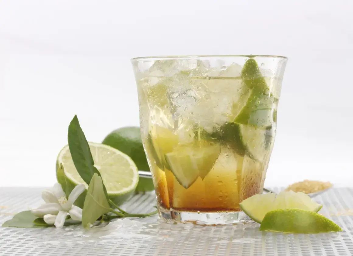 Orzeźwiająca caipirinha przepis: drink z lodem, limonką i cukrem. Obok leżą świeże limonki i kwiatki.