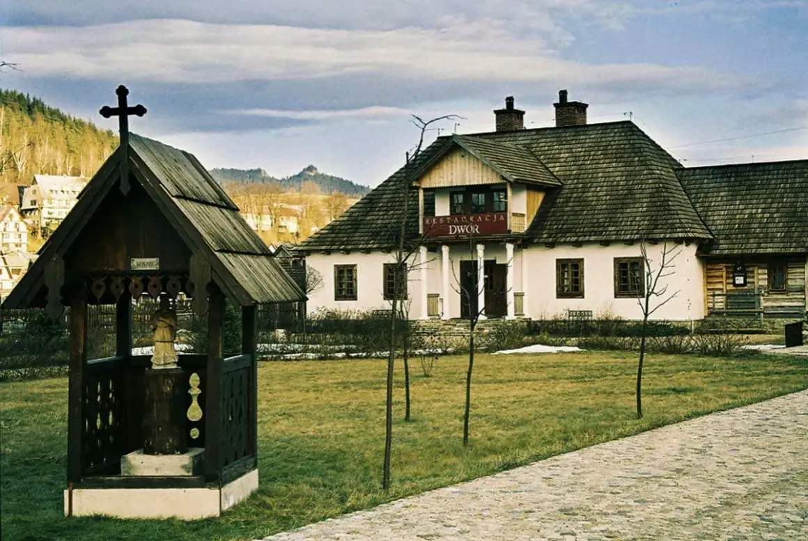 Drewniana kapliczka z krzyżem i figurą, obok restauracja "Dwór". W tle sosny i zabudowania. Idealne miejsce na camping.