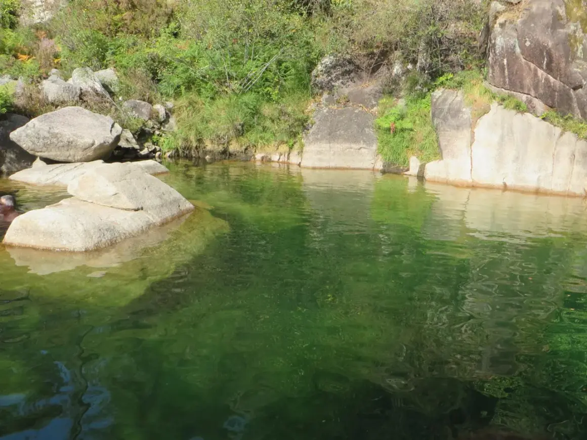 Un hombre se baña en la poza de agua verde de la corga da fecha, rodeada de rocas y vegetación.