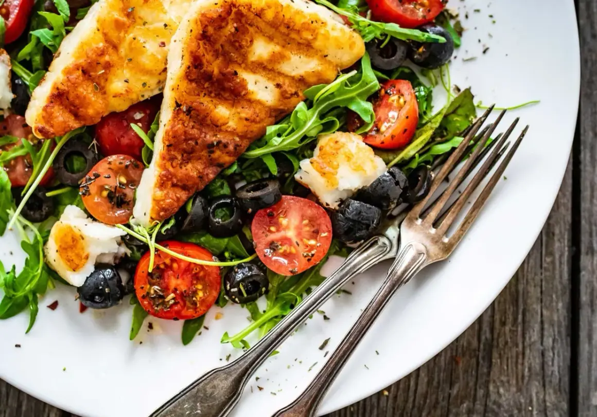 Pyszna sałatka z grillowanym halloumi, pomidorkami koktajlowymi i czarnymi oliwkami na rukoli. Idealna na lekki posiłek.