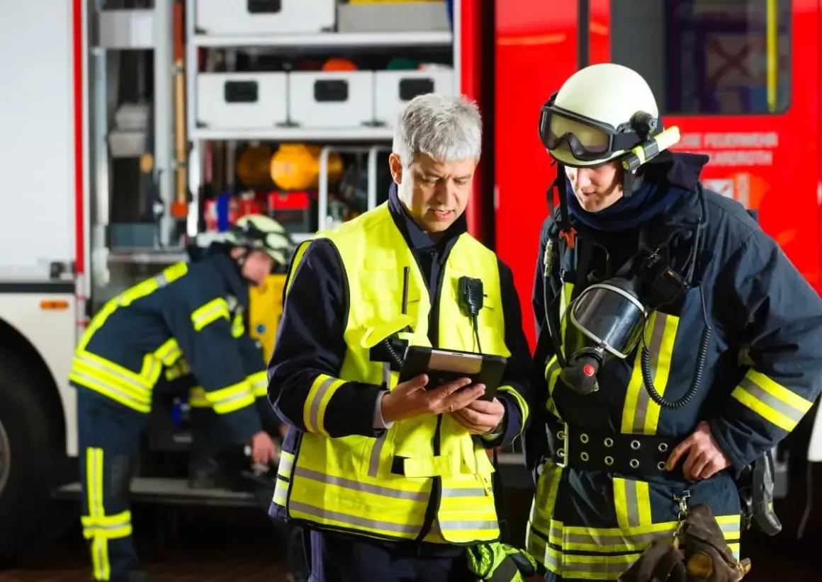 Feuerwehrleute besprechen Einsatzdetails. Ein Mann in Warnweste prüft ein Tablet, während ein Kollege in voller Montur mit Atemschutzmaske daneben steht. UVV Feuerwehr wird beachtet.