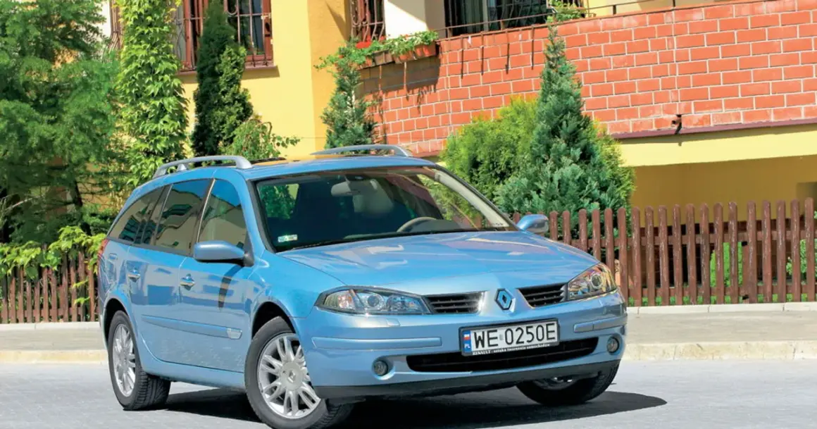 Błękitne kombi Renault Laguna 2 z relingami dachowymi stoi przed domem z żółtą elewacją i drewnianym płotem.