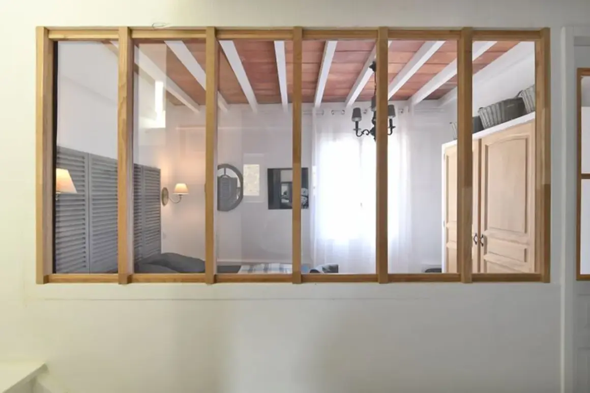 Une verrière en bois sépare deux pièces. On voit des volets, un lit, un lustre et des portes. Idéal pour savoir comment installer une verrière.