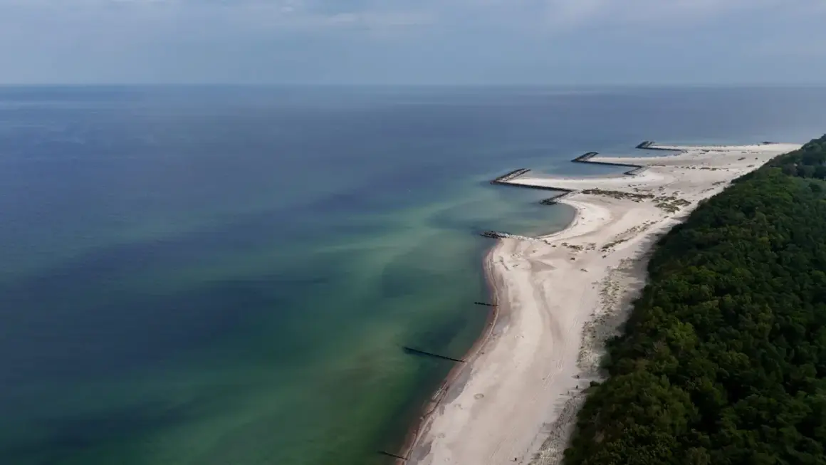 Plaża w Jarosławcu, zwana czasem "plażą dubaj", z kamiennymi falochronami i zielonym lasem.