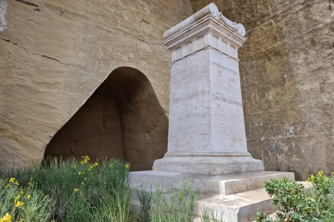 Monumento a Giacomo Leopardi nel Parco Virgiliano a Piedigrotta, con un'antica grotta sullo sfondo e fiori gialli.
