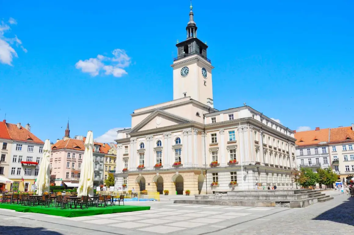 Rynek w mieście nad Prosną. Ratusz z wieżą zegarową góruje nad placem, gdzie stoją stoliki kawiarniane.