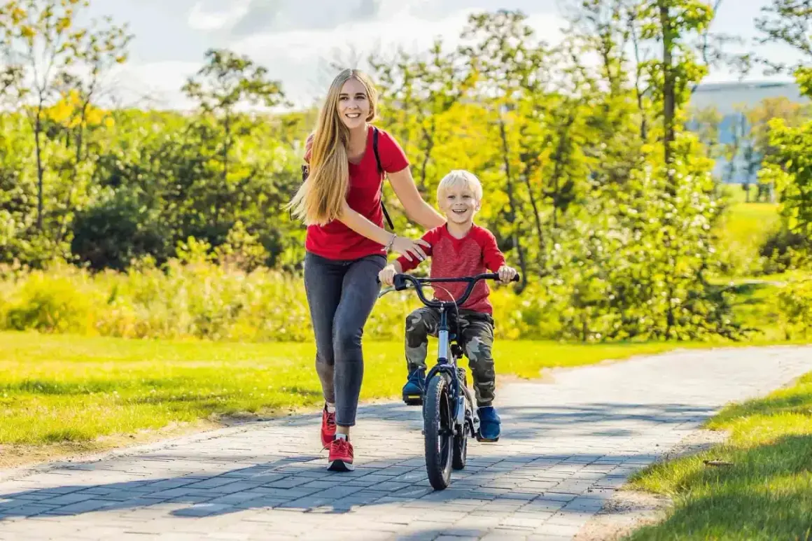 Mama pomaga synkowi, jak nauczyć dziecko jazdy na rowerze. Uśmiechnięci, na ścieżce w parku, wśród zieleni.