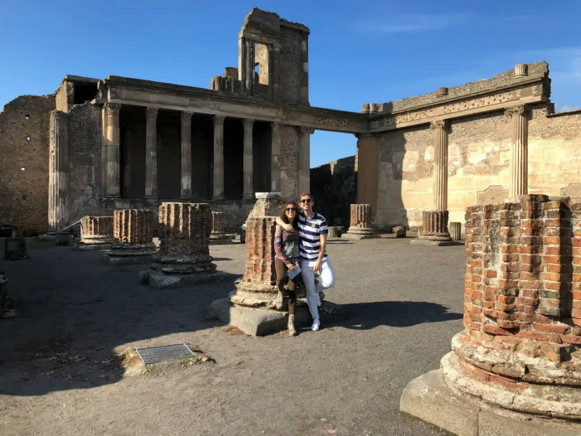 Pareja posa frente a ruinas antiguas, preguntándose donde está Pompeya. El sol ilumina columnas rotas y edificios históricos.