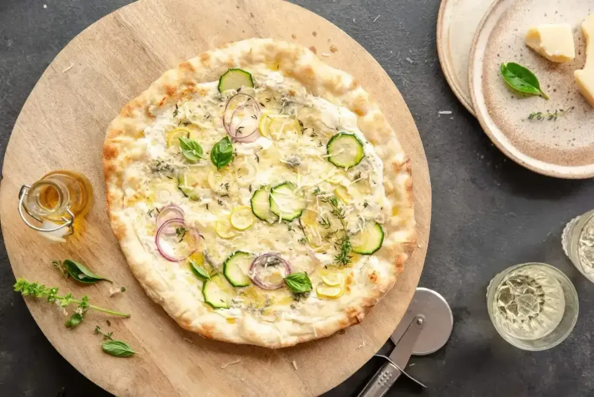 Pyszna pizza bez sosu pomidorowego z cukinią, cebulą i ziołami, podana na drewnianej desce.