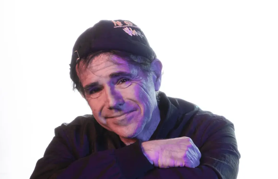 Andrea Satta, con un sorriso enigmatico, indossa un cappellino scuro e una felpa. La luce colorata crea un'atmosfera suggestiva.
