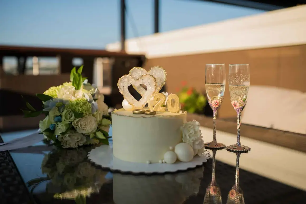 Torta per 20 anni di matrimonio con cuori, anelli e numero 20 dorato. Due flute di champagne decorate e un bouquet di fiori completano la scena.