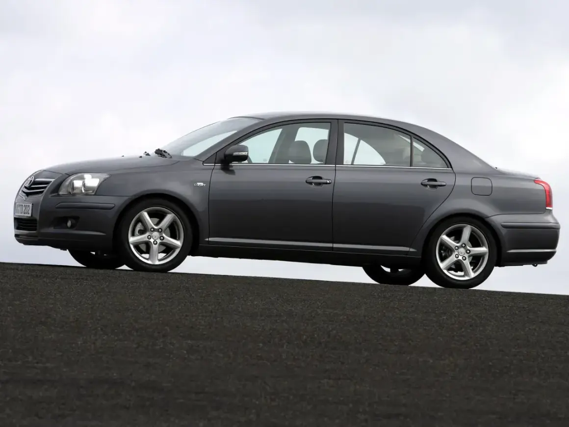 Szary sedan Toyota Avensis 1.8 benzyna stoi na ciemnej nawierzchni, z niebieskim niebem w tle.