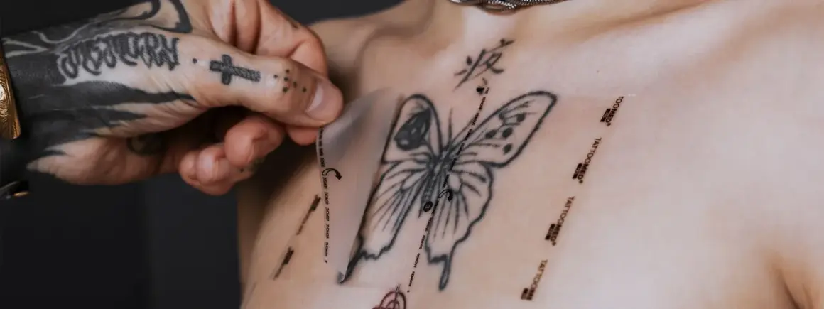 Ein Mann mit tätowierter Hand zieht eine Schutzfolie von einem frisch gestochenen Schmetterling-Tattoo auf der Brust ab. Pflege danach ist wichtig.