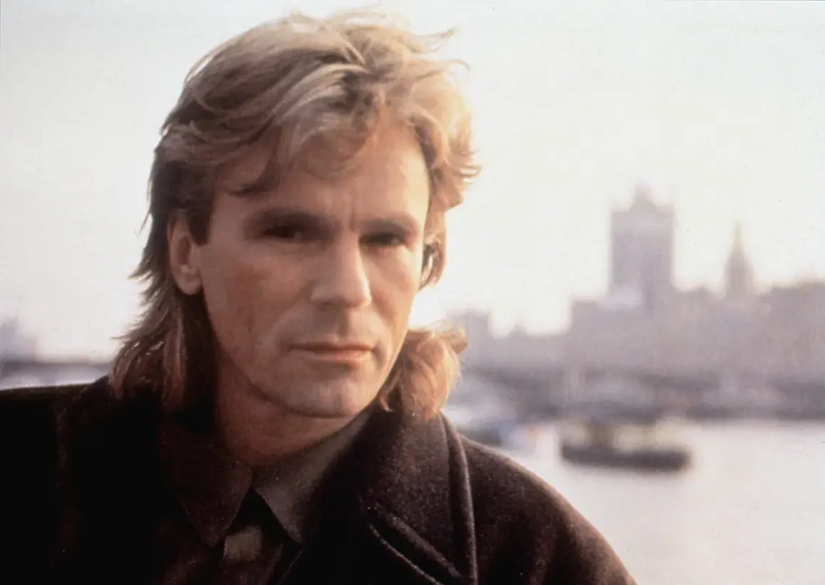 MacGyver obsada: Richard Dean Anderson jako MacGyver, z charakterystyczną fryzurą i w płaszczu, na tle mglistego miasta.