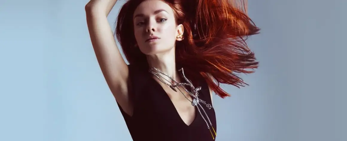 Mujer con cabello cobrizo vibrante, moviéndose con gracia. Luce un collar plateado llamativo y un pendiente dorado.