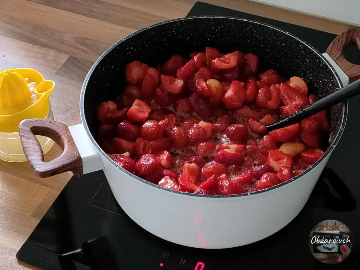 Duży garnek z gotującymi się truskawkami na kuchence indukcyjnej. Obok żółta wyciskarka do cytrusów.