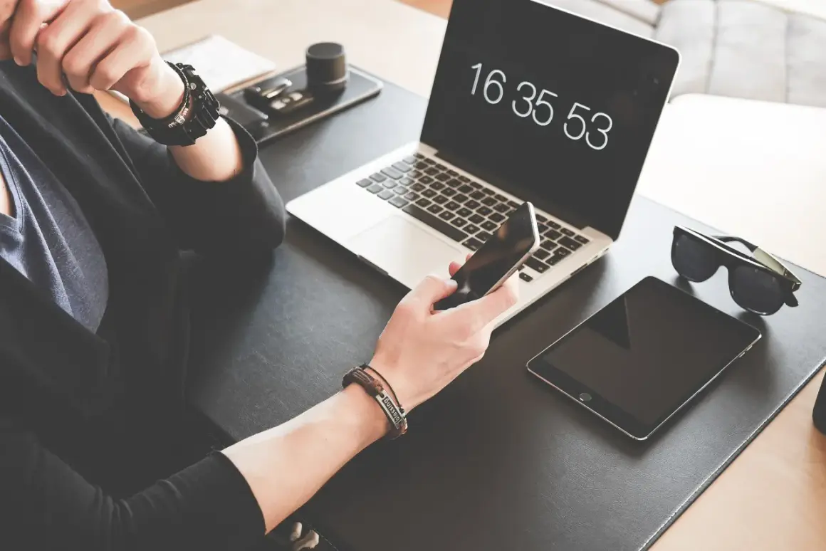 Osoba trzyma iPhone'a, obok leży tablet i laptop z widocznym zegarem.
