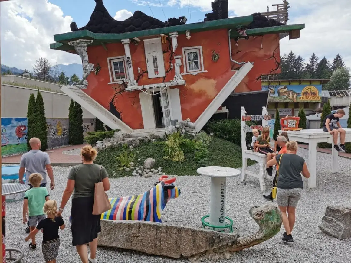 Ein rotes Haus steht auf dem Kopf, die Treppe führt ins Nichts. Eine Familie bestaunt das Spektakel, bunte Skulpturen und ein Dino-Land im Hintergrund.