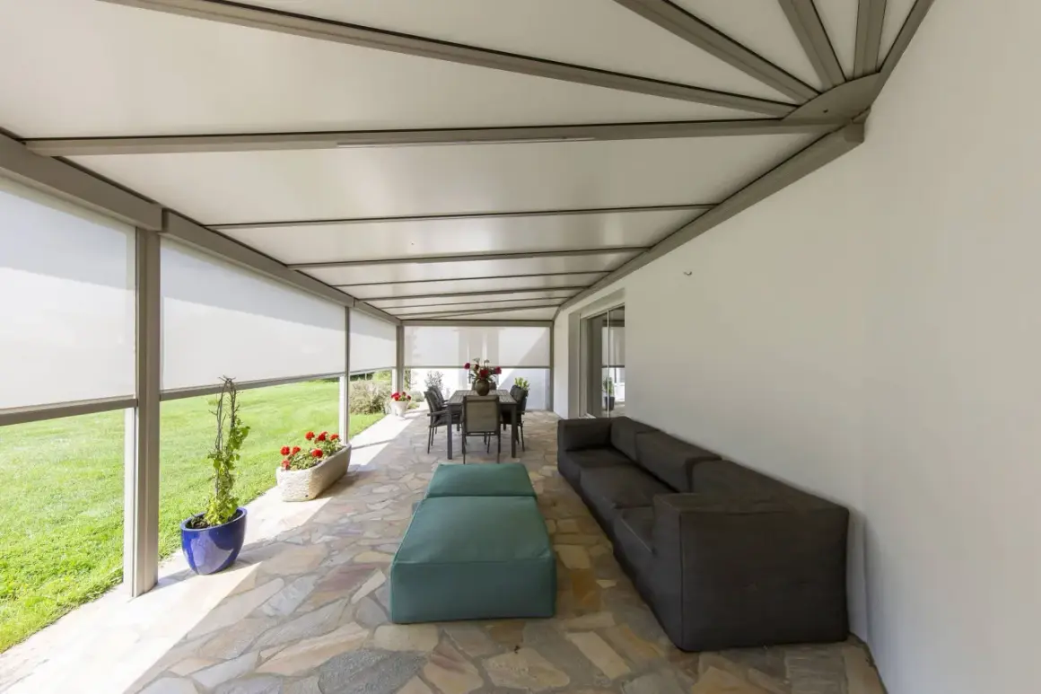 Telo per pergolato fissato con sistema a scomparsa, creando un'area relax con divano e tavolo da pranzo.