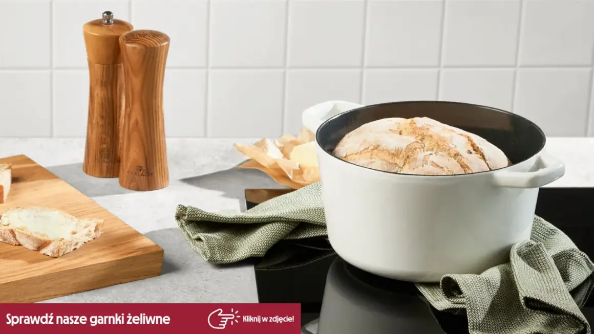 Biały garnek żeliwny z upieczonym chlebem, obok deska z masłem i młynki do przypraw.