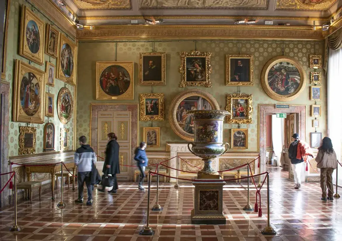 Interno di una sala del Palazzo Pitti, con opere d'arte alle pareti, un grande vaso e visitatori.