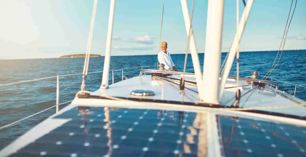 Un uomo sorride sul ponte di una barca a vela, con un impianto pannello solare che riflette il cielo azzurro.