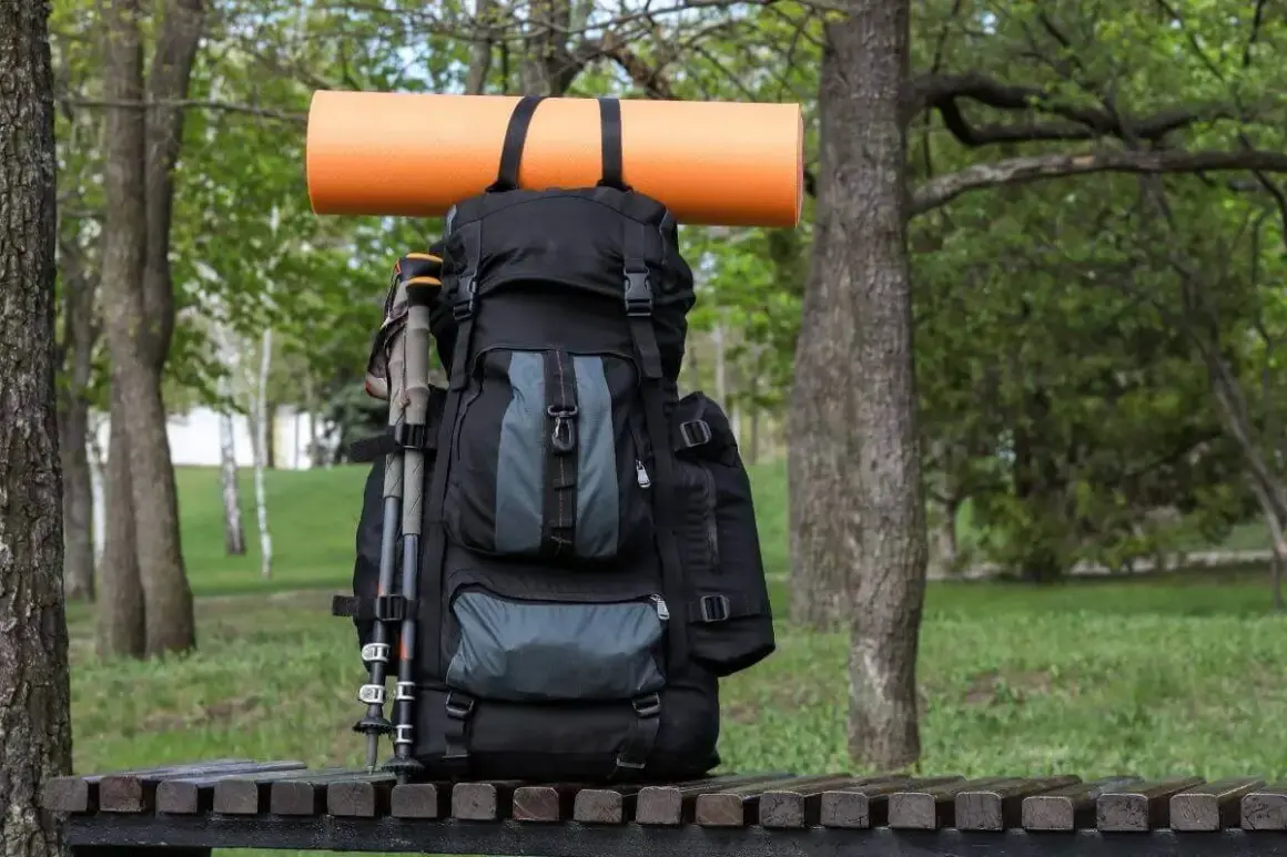 Ein schwarzer Rucksack mit Wanderstöcken und einer orangefarbenen Isomatte, die mit Schlaufen befestigt ist, steht auf einer Holzbank im Grünen.