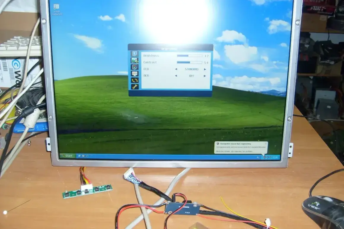 Na biurku leży monitor z pulpitem Windows XP, obok kable i płytka sterująca. To może być pomocne, jak podłączyć monitor do laptopa.