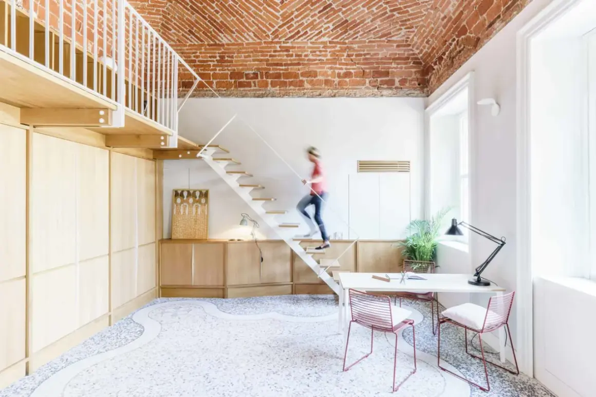 Soggiorno con soppalco a vista, soffitto in mattoni, scala bianca e una persona che sale. Arredamento moderno e minimalista.
